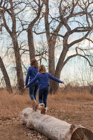 Two people in matching blue outfits balance while walking on a fallen log in a winter forest landscape.