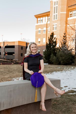 A person in a black top and purple skirt sits casually on a concrete ledge near a brick building at sunset.