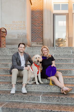 A couple sits on granite steps with their fluffy white dog wearing a red collar and leash.