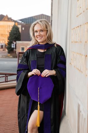 Graduate wearing purple and black academic regalia poses for graduation photos on campus.