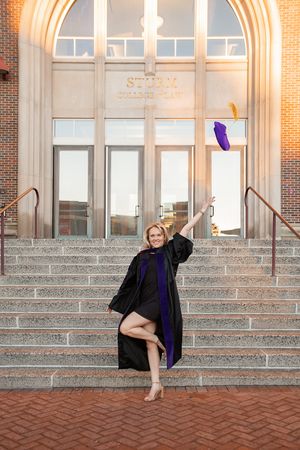 A graduate in a black robe poses joyfully on the steps of a brick building with arched windows.