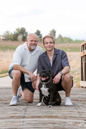 Two men and a black and white dog pose together on a wooden boardwalk with a natural landscape behind them.