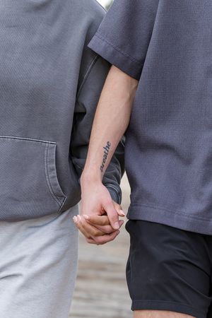 Two people holding hands, one with a breathe tattoo on their forearm, both wearing casual gray outfits.