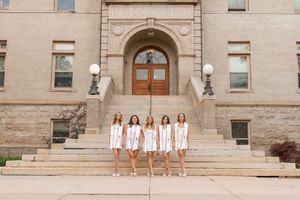 Six women in white dresses pose on steps of a historic stone building with arched wooden doors.