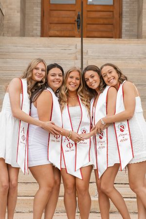Five Alpha Phi sorority sisters in white dresses posing with graduation stoles on stone steps.
