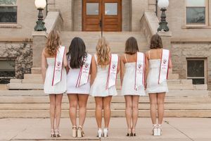 Five women in white dresses showing the backs of their Alpha Phi graduation stoles on steps.