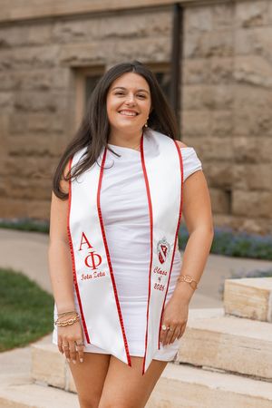 Smiling woman in white dress wearing Alpha Phi Iota Zeta Class of 2026 graduation stole outdoors.