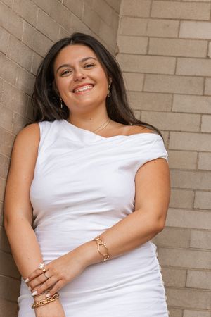 Smiling young woman in white off-shoulder dress with gold jewelry leaning against brick wall.