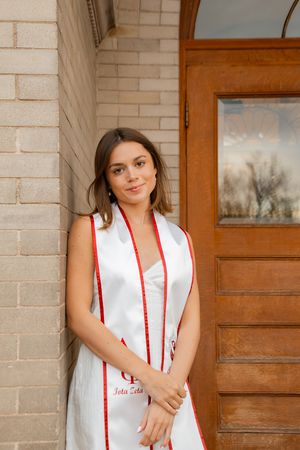 Young woman in graduation stole with red trim leaning against brick wall near wooden door.