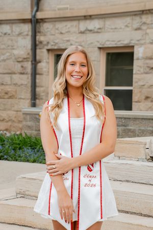Smiling blonde woman in white dress wearing Alpha Phi Class of 2026 graduation stole outdoors.
