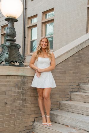 Young blonde woman smiling in white mini dress and heels on stone steps near ornate lamp post.
