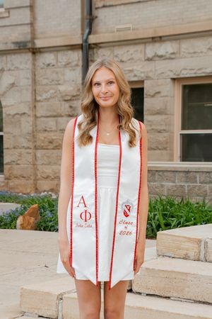 Young woman in white dress wearing Alpha Phi graduation stole, Class of 2026, posing outdoors near stone building.