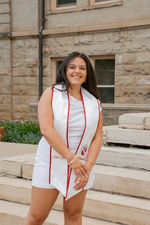 Smiling graduate in white dress and red-trimmed stole posing on stone steps outdoors.