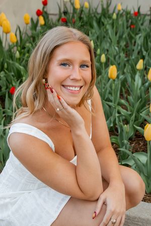 Smiling blonde woman with blue eyes posing by colorful tulips in white dress with red manicure.