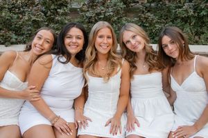 Five young women in white dresses smiling together outdoors against a green ivy wall.