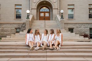 Group of six young women in white outfits sitting on steps of a classic stone building.