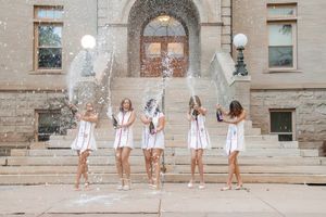 Five graduates in white dresses celebrating with champagne spray on university building steps.