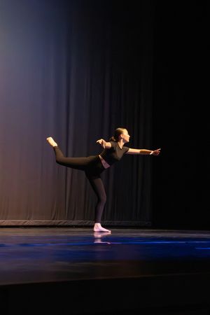 A dancer in black performs a graceful horizontal pose on a dark stage with blue lighting.