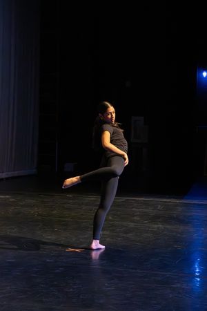 A dancer performs a standing pose on a dark stage with blue lighting, wearing black dance attire.