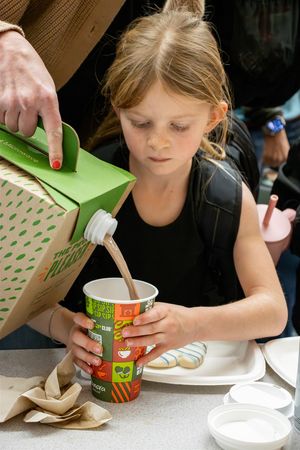A child eats from a disposable cup with a spoon while sitting at a table with brown paper bags nearby.