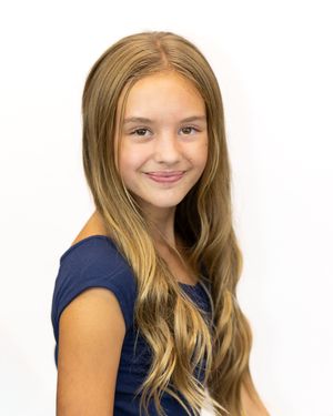 Young girl with long wavy blonde highlighted hair smiling, wearing a navy blue top against a white background.