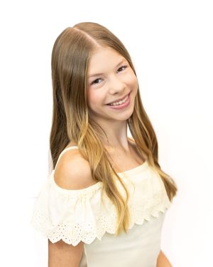 Smiling young girl with long blonde hair wearing a white off-shoulder top, posing for a portrait photo.