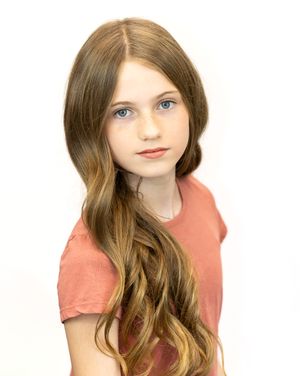 Young girl with long wavy brown hair and blue eyes wearing a pink shirt, posing for a portrait photo.