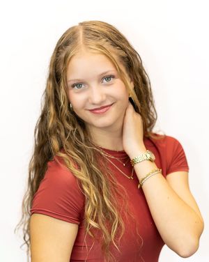 Young teen girl with long wavy blonde hair wearing a red top and gold jewelry smiles for a portrait.
