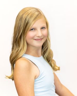 Smiling young girl with long blonde hair wearing a light blue sleeveless top against a white background.