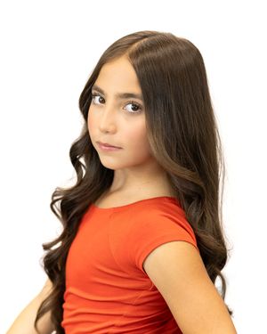 Young girl with long wavy dark brown hair wearing a red top, posing against a white background.