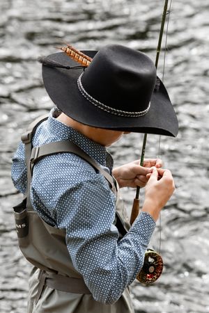 Fly fisherman in black cowboy hat and waders carefully ties fishing line on rod near rippling river.