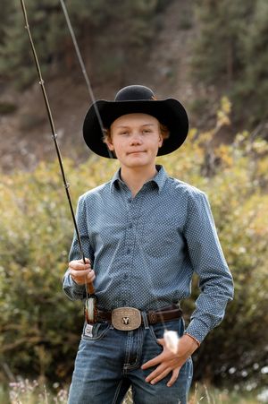 Young cowboy in black hat and western shirt holds fishing rod outdoors near trees.