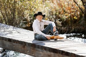 Young man in cowboy hat and boots sitting relaxed on a wooden bridge surrounded by autumn trees.