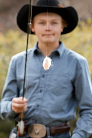 A young boy in a black cowboy hat holds a fishing rod with a fly lure in focus in the foreground.