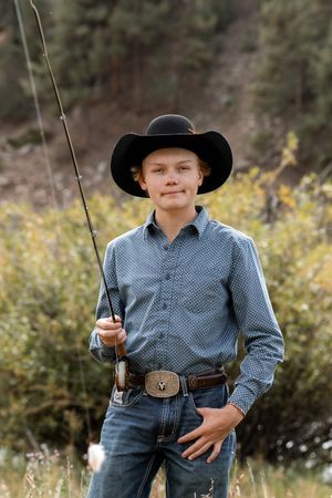 A young boy in a black cowboy hat and blue shirt holds a fishing rod outdoors near trees and brush.