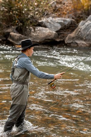 A fly fisherman wearing a cowboy hat and waders casts a line while standing in a rocky mountain river.