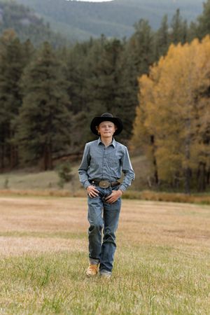 Young boy in cowboy hat, blue shirt, jeans, and belt walks through a field with autumn trees and mountains behind him.