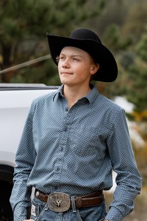 Teen boy wearing black cowboy hat and western shirt with decorative belt buckle standing outdoors.