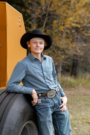 Young boy in black cowboy hat and western shirt leaning against large tractor tire outdoors in autumn.