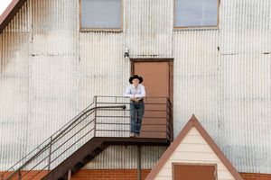 Person in cowboy hat standing on metal staircase landing of rustic corrugated metal building exterior.