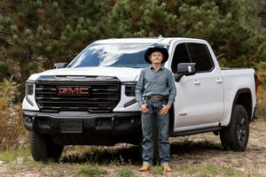 Man in cowboy hat standing beside a white GMC Sierra pickup truck outdoors near pine trees.