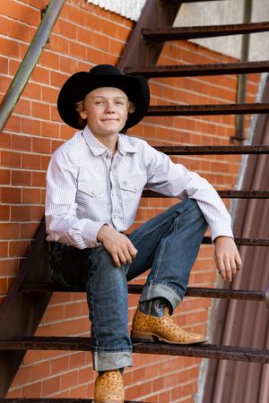 Young boy in cowboy hat, jeans, and exotic boots sitting on metal stairs against brick wall.