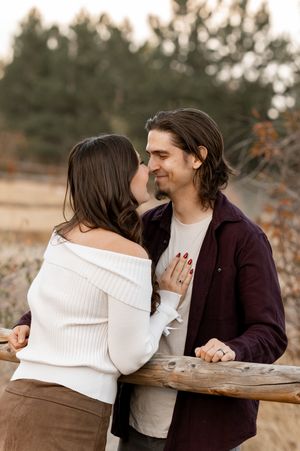 A couple shares a romantic moment outdoors, leaning against a wooden fence in autumn.