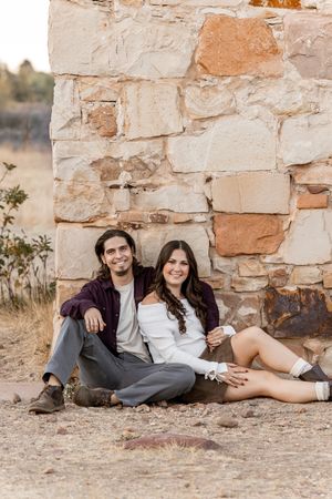 A smiling couple sits together against a rustic stone wall outdoors, dressed in casual fall attire.