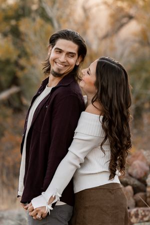 A smiling man in a burgundy shirt holds hands with a woman in a white sweater outdoors in fall.