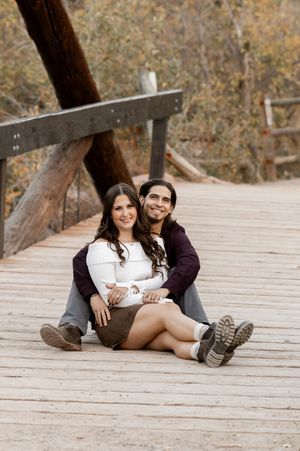 Couple sitting on a wooden bridge in autumn, smiling and embracing in a cozy outdoor engagement photo.