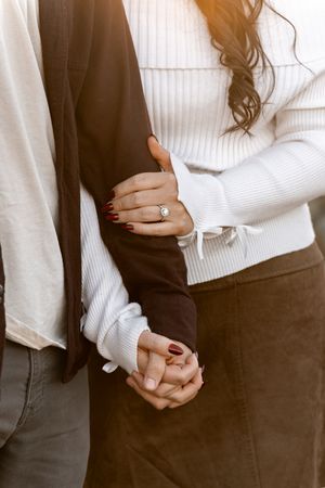 Couple holding hands, woman wearing engagement ring with red nails, dressed in white sweater and brown skirt.