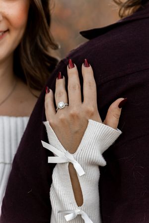 Close-up of woman's hand displaying a diamond halo engagement ring with dark red nails, resting on a burgundy jacket sleeve.