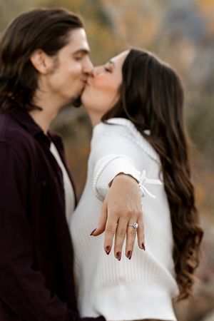 Couple kissing outdoors while showing off engagement ring with diamond solitaire, wearing white sweater and burgundy shirt.