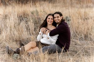 Couple sitting together in a golden grass field, embracing and smiling during an outdoor engagement photo session.